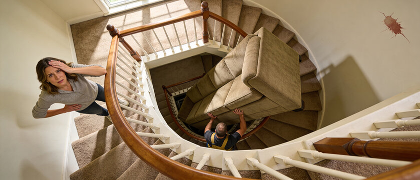 Woman watching a man struggling to move a large sofa down a winding staircase in apartment move. Difficult home moving process concept.