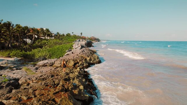 Woman walks on rocky shore as waves crash with turquoise water beside palm trees in Tulum Mexico