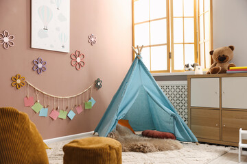 Colorful paper notes and beautiful decor hanging on beige wall near toy wigwam in stylish children's room with chest of drawers and poufs. Interior design © New Africa