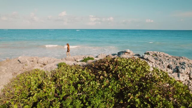 Woman walking along rocky shore by turquoise ocean waters in Tulum, Mexico
