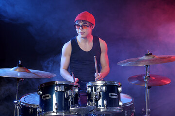 Obraz premium Teenage boy in glasses and hat learning how to play drums in color lights and smoke against dark background
