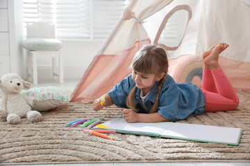 Little girl drawing in sketchbook near toy wigwam at home © New Africa