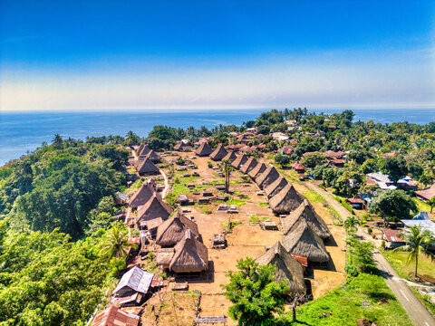 Aerial view of traditional thatched-roof houses at Kampung Adat Nggela surrounded by lush tropical greenery and the blue sea in Wolojita, East Nusa Tenggara, Indonesia.