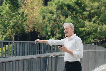Fototapeta premium Older man standing outdoors holding smartphone and leaning on railing with green trees in background, thoughtful expression on face