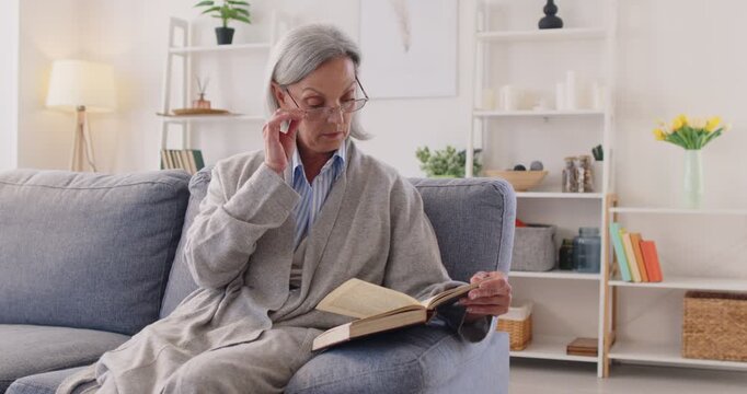 Senior woman enjoys reading a book at home, engaging in a peaceful leisure activity. Love for literature and relaxation creates a comforting hobby within personal space, like a small library at home.