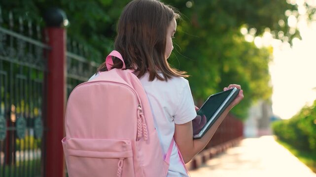 Teenage girl walks down street with tablet in hand. Student hurries to school lesson city street carrying digital tablet. Child with backpack on back hurries to school. Children education concept