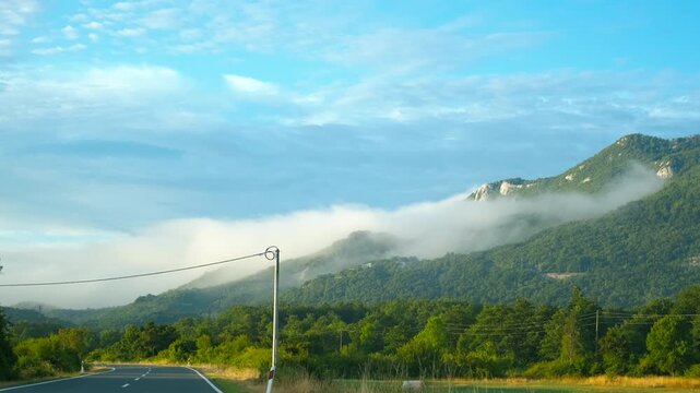 Driving along a foggy mountain road at sunrise. Scenic view from a moving car window of a winding mountain road and lush green forest, with a beautiful morning fog enveloping the hillsides