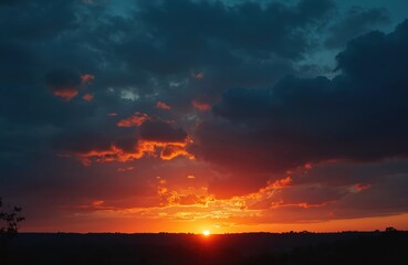 Fototapeta premium Dramatic dark sunset sky with bright orange sun low on horizon. Clouds glow with warm fiery light over dark land silhouette. Natural beauty.