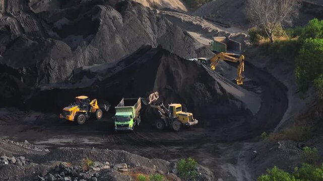 Heavy machinery including excavators and trucks is actively digging and transporting dark volcanic sand, creating large industrial piles within the Amed quarry in Bali. Aerial drone flight footage