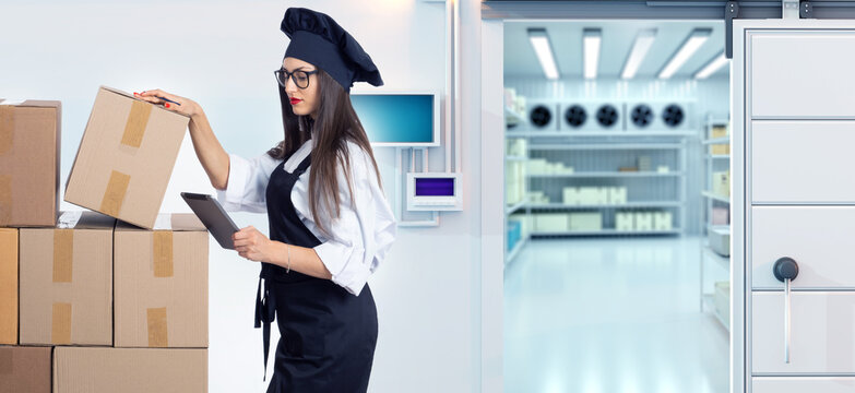 Restaurant cold storage. Chef checks boxed food supplies with a tablet and organizes inventory near a refrigerated warehouse room.