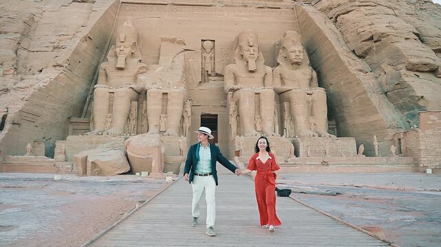 american young man traveler wearing white hat with his wife with red dress exploring ancient Egyptian abu simble temple with background of ramsses ii statues. Egypt.