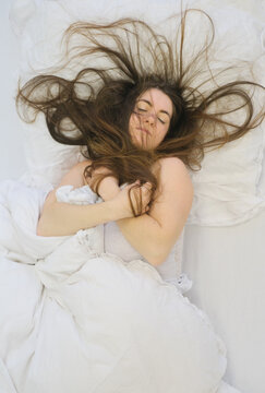 Young woman sleeps on white bed with long hair spread in dynamic strands across pillow holding blanket close creating contrast between motion and restful peaceful atmosphere