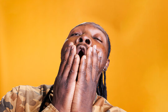 Portrait of exhausted african american person covering mouth with hand being tired while posing in studio standing over yellow background. Man yawning having overworked expression