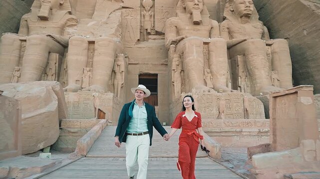 american young man traveler wearing white hat with his wife with red dress exploring ancient Egyptian abu simble temple with background of ramsses ii statues. Egypt.