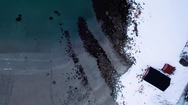 Drohne fliegt nach unten &uuml;ber arktischen Strand mit Fischerhaus und Wellenbrechern in Nordnorwegen