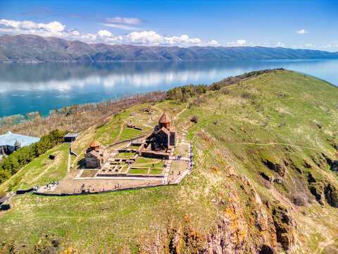 Aerial view of the historic Sevanavank monastery on a green peninsula with people walking on paths overlooking the blue waters of Lake Sevan in Sevan, Gegharkunik Province, Armenia.