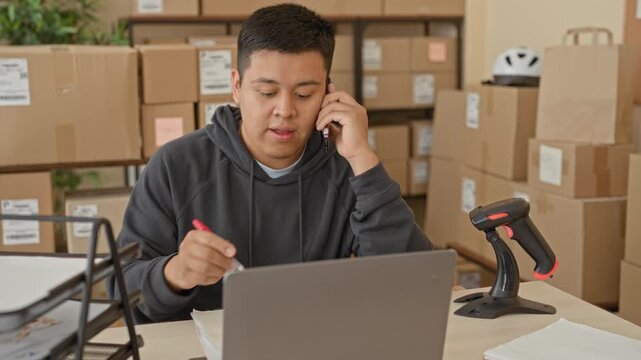 Man holds smartphone to ear and writes by laptop amid stacked parcel boxes and barcode scanner in a building; small business stress.