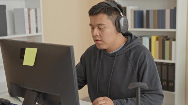 Man wearing headphones types at computer while holding a pen and leaning toward the monitor in a building office with bookshelves; focus productivity.
