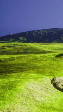 A large green field stretches under a clear sky. Mountains are visible in the background. The light from the setting sun creates a gentle glow. Small rocks are scattered throughout the field.