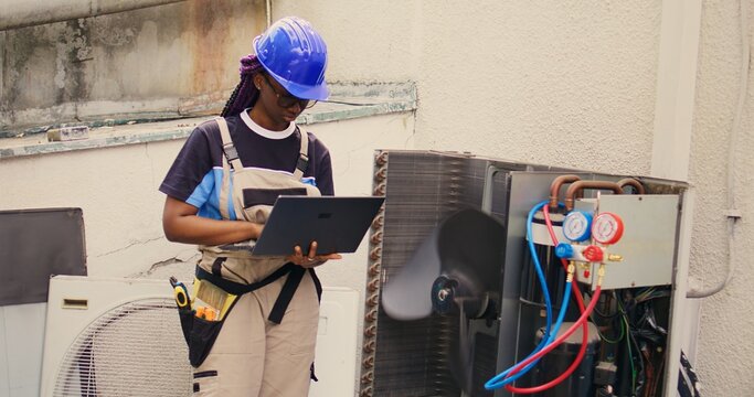 Qualified african american electrician working on outside hvac system, holding laptop. Licensed serviceman optimizing external air conditioner performance, ensuring it operates at maximum capacity