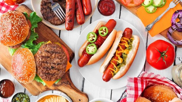 Summer BBQ food table scene with hot dog and hamburger buffet. Slow rotating zoom motion. Overhead view on a white wood background.