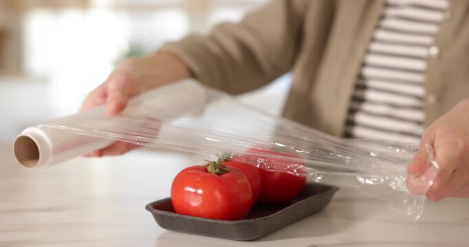 Woman wrapping container with fresh tomatoes in cling film at light table, closeup