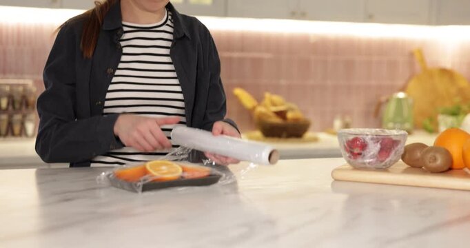 Woman wrapping container with fresh orange slices in cling film at light marble table, closeup. Camera moving