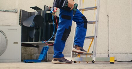 Meticulous mechanic in professional uniform with technical equipment climbing folding ladder to do maintenance on rooftop hvac system. Professional worker commissioned to check up on air conditioner © DC Studio