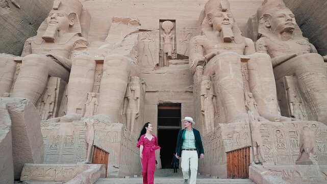 american young man traveler wearing white hat with his wife with red dress exploring ancient Egyptian abu simble temple with background of ramsses ii statues. Egypt.