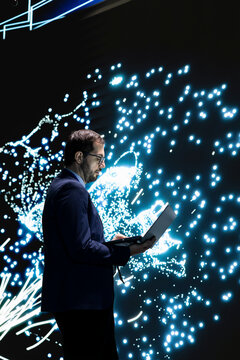 Technician monitors deep learning AI model patterns using laptop. RD lab employee studies artificial intelligence neural network algorithm nodes on digital wall to understand processing using notebook