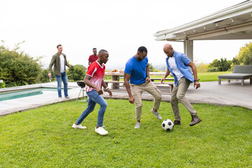 Obraz premium Diverse male friends kicking soccer ball on backyard lawn beside picnic table and charcoal grill