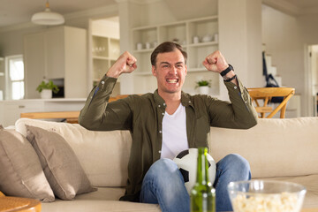 Adult man holding soccer ball on sofa with popcorn, reacting to TV goal, raising fists