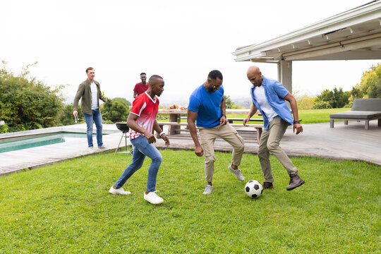 Diverse male friends kicking soccer ball on backyard lawn beside picnic table and charcoal grill