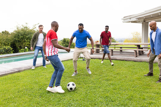 Diverse male friends playing with soccer ball on grassy lawn beside house with pool and speaker