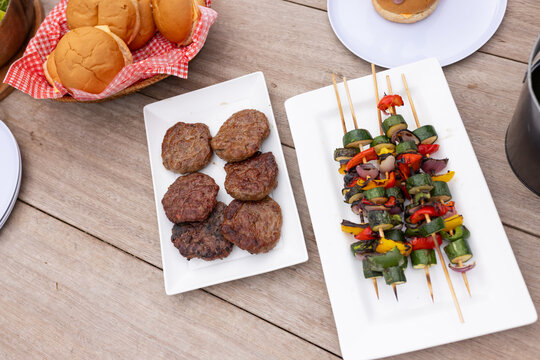 White plates sitting on picnic table holding six burger patties, vegetable skewers and bread basket