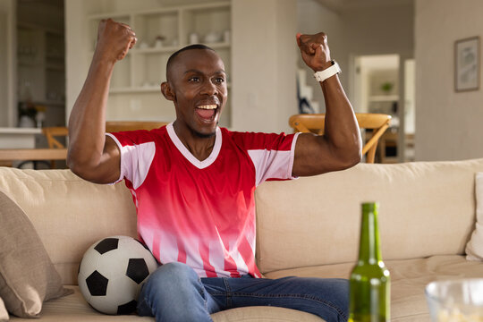 Male cheering and raising fists while sitting in living room wearing red jersey with soccer ball