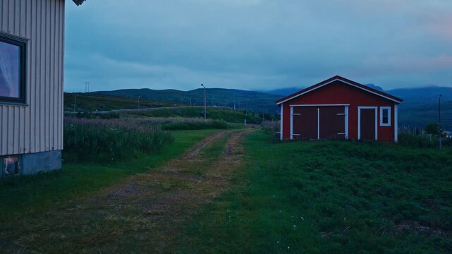 Kokelv, Hammerfest, Finnmark, Norway - A Narrow Dirt Path Leads Between a Wooden House and a Red Shed, With Rolling Hills in the Distance - Drone Flying Forward