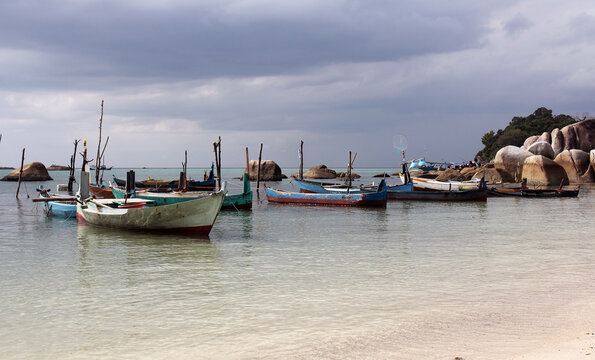The image features a collection of fishing boats resting in the shallow water near a beach with large rock formations.