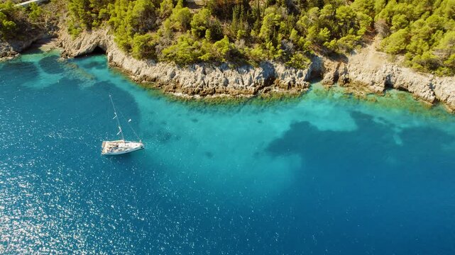 White sailboat anchored in a turquoise bay near a rocky forested coast in summer