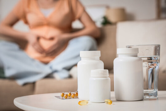 Folic acid pills with glass of water on table against pregnant woman sitting at home, closeup