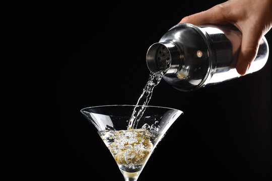 Female bartender pouring tasty martini cocktail into glass with olives on dark background
