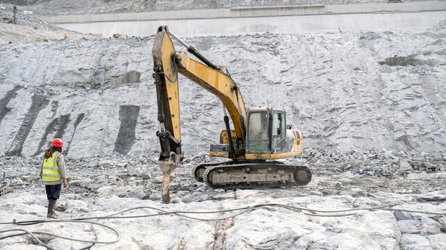 Excavator breaking rock at dam construction site with worker supervising.
