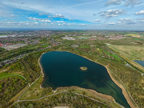 Aerial view of Albert Village Lake with its small central island, surrounded by lush green forests and the urban landscape under a blue sky in Swadlincote, England, United Kingdom.