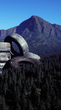 A pile of discarded tires sits on the ground among tall trees in a mountainous area. The clear sky shows the surrounding peaks under bright sunlight.