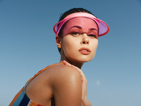 Close-up portrait of a young woman wearing a pink visor, sunlit skin and glossy lips, looking confidently toward the camera with a bold summer vibe and radiant glow.