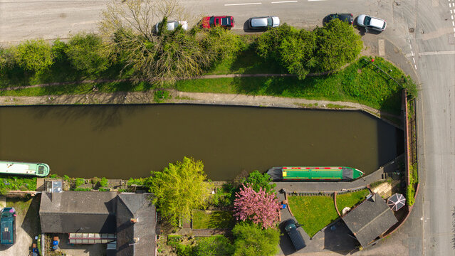 Aerial view of narrowboats moored on a canal surrounded by spring blossom, lush green trees, and residential houses in Barton-under-Needwood, England, United Kingdom.