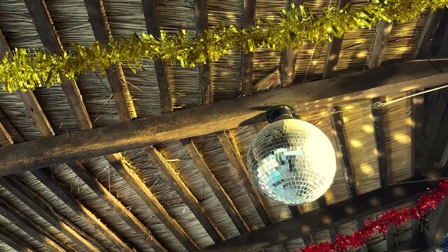 Shiny disco ball hanging from rustic wooden beam under thatched roof with festive christmas decorations.
