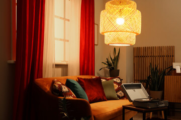 Comfortable sofa with modern laptop and glowing lamp near red curtains in living room