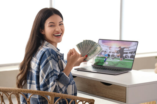 Happy young woman with money and laptop after winning her sports bet at home