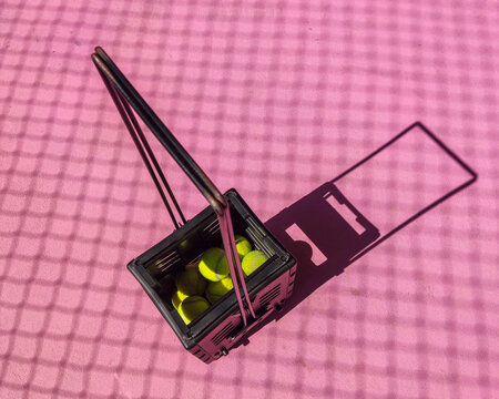 Tennis ball hopper placed at center of court under bright daylight, showcasing sports training equipment used for collecting balls during practice sessions on outdoor tennis field.